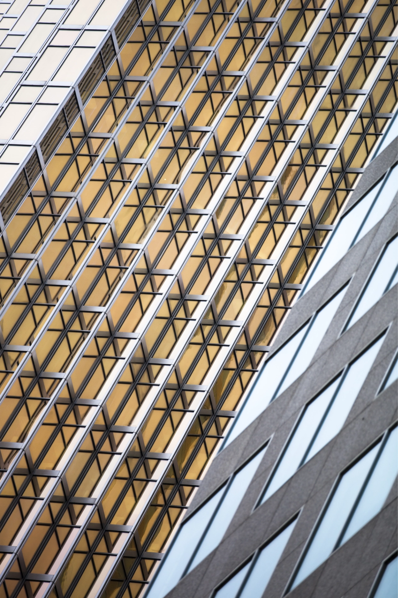 Modern bir binanın cam cephe detayını gösteren, çapraz çizgiler oluşturan metal çerçeveler ve altın tonlu yansımalarla geometrik bir mimari görünüm.