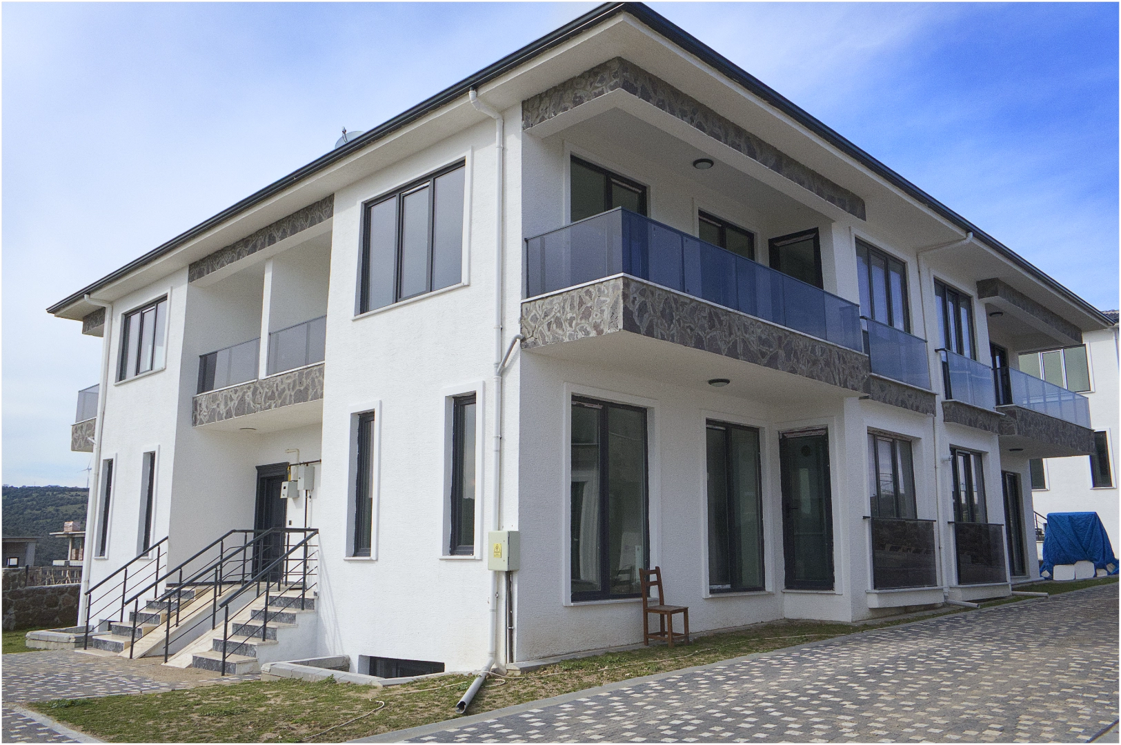 Modern mimariye sahip, beyaz cepheli iki katlı konut binası; cam balkon korkulukları, geniş pencereler ve taş kaplama detaylarıyla döşeli avlu içinde yer alıyor.