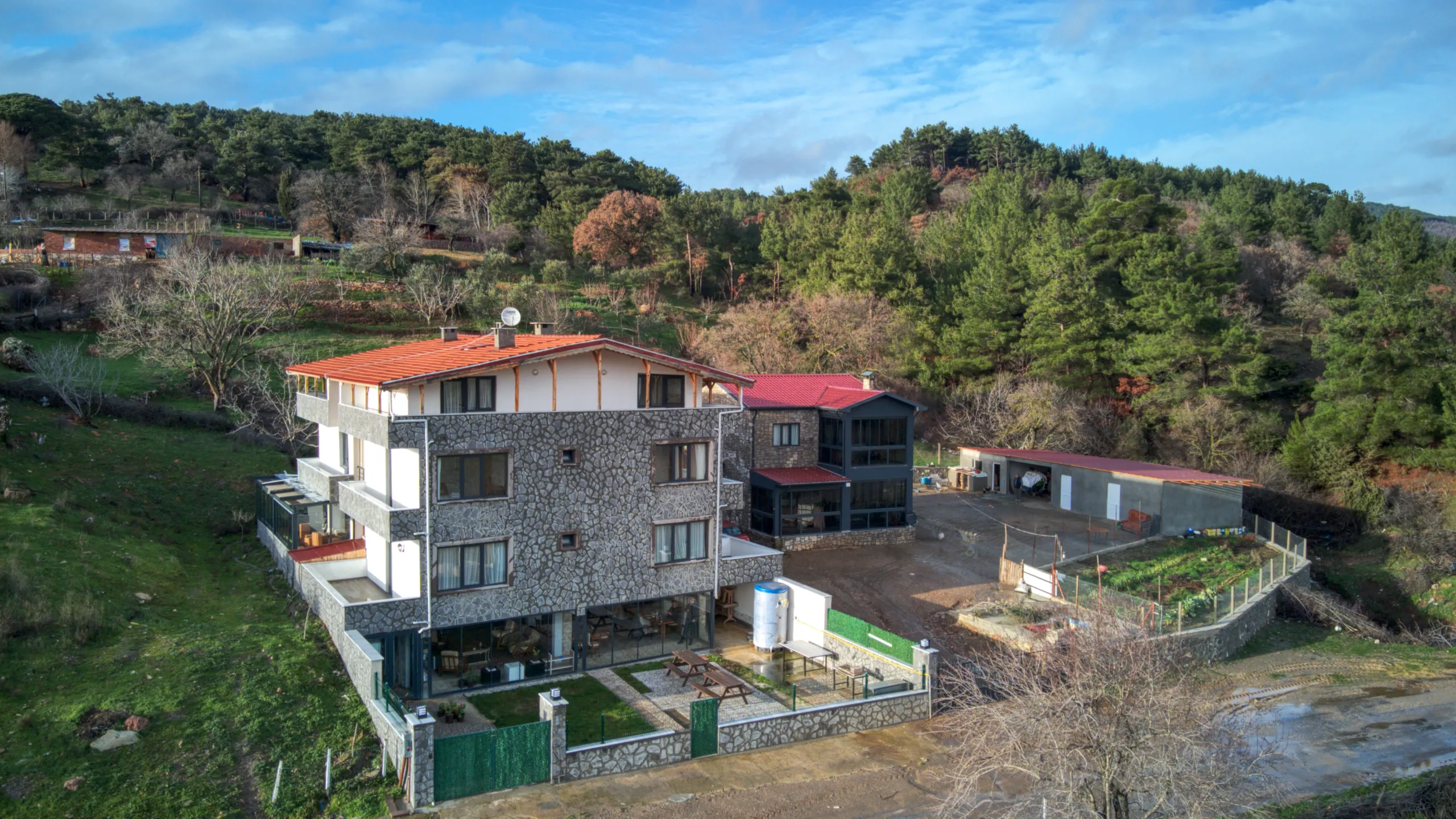 Ormanla çevrili kırsal bir alanda yer alan, taş kaplamalı dış cepheye ve kırmızı çatıya sahip çok katlı müstakil bir evin havadan görünümü. Evin yanında ek yapılar, çevresinde bahçe alanları ve ağaçlarla kaplı yamaçlar görülüyor.