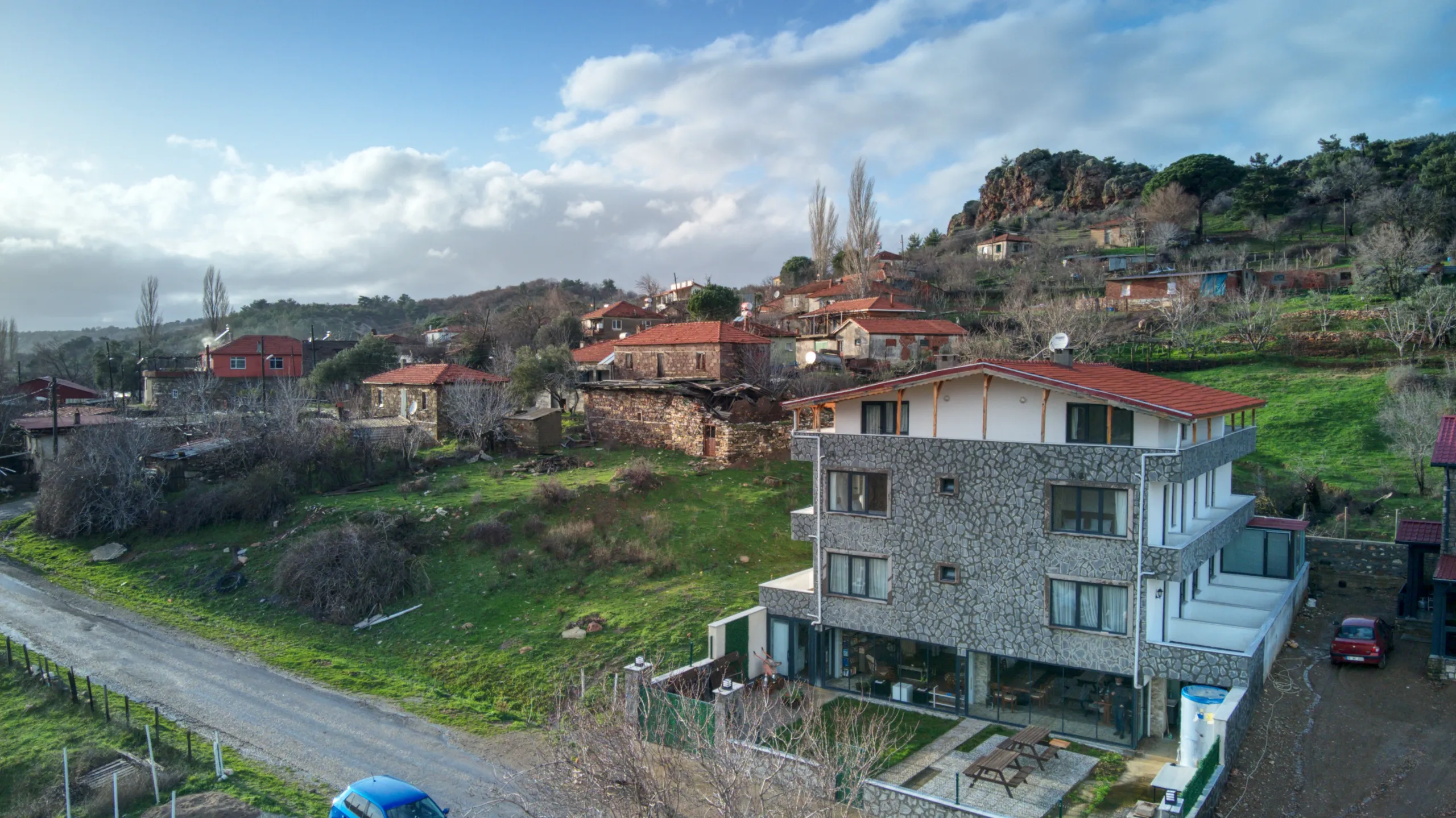 Kırsal bir yerleşim alanında bulunan, taş kaplamalı dış cepheye ve kırmızı çatıya sahip çok katlı bir evin havadan görünümü. Evin çevresinde yeşil arazi, köy evleri, dar bir yol ve arka planda kayalık tepeler ile bulutlu gökyüzü yer alıyor.