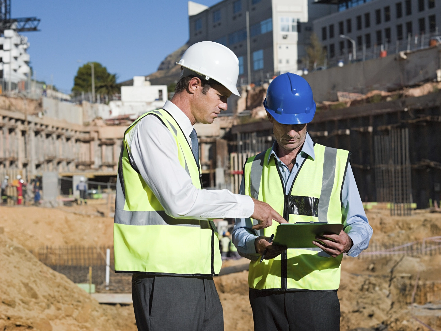Şantiye sahasında baret ve reflektörlü yelek giyen iki mühendis, inşaat uygulama sürecini takip ederek proje ilerleyişini tablet üzerinden kontrol ediyor.