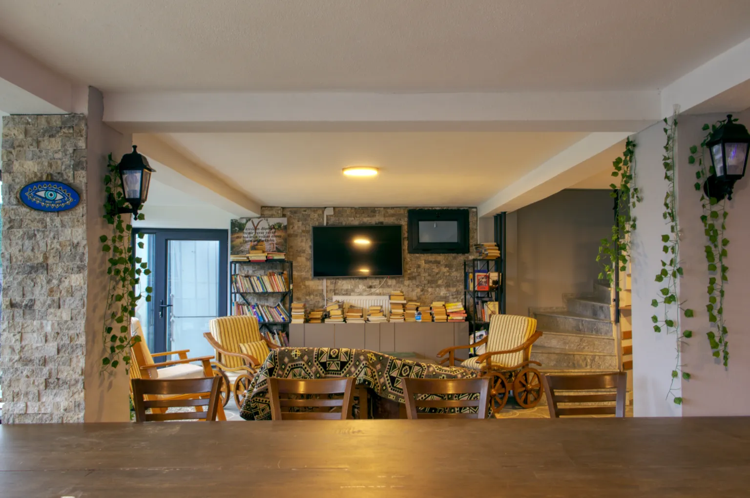 Baharlar Otel’in oturma alanını ve merdiven bölümünü gösteren, koltuklar, kitaplıklar ve taş duvar detayları içeren iç mekân görünümü.