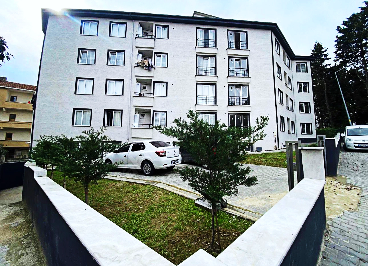 Siyah pencere detaylarına sahip çok katlı beyaz bir apartman, önünde park halinde beyaz bir otomobil ve küçük ağaçların bulunduğu çimenlik bir bahçe.