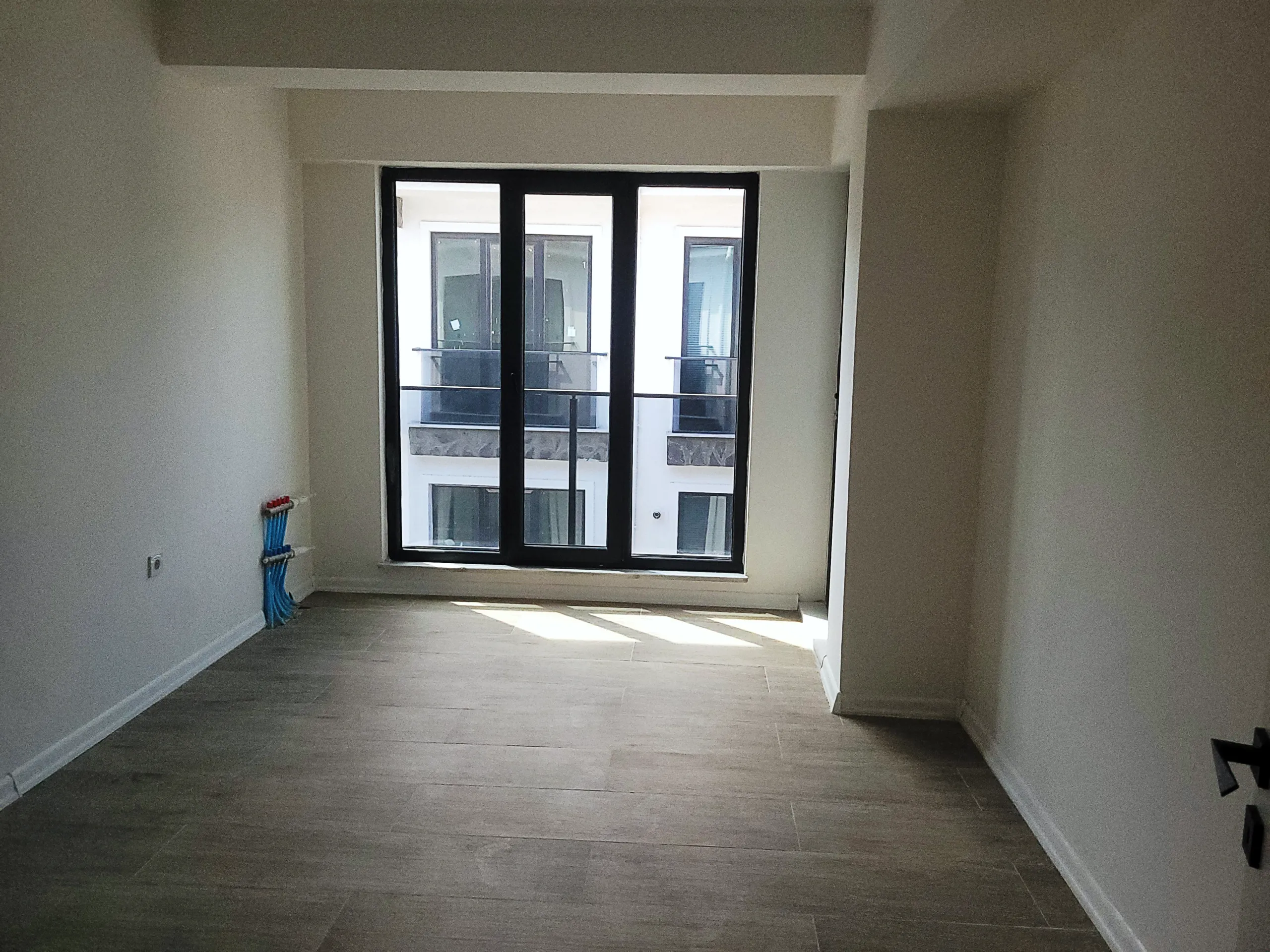 Empty living space with grey flooring and natural light entering through a wide black-framed window.
