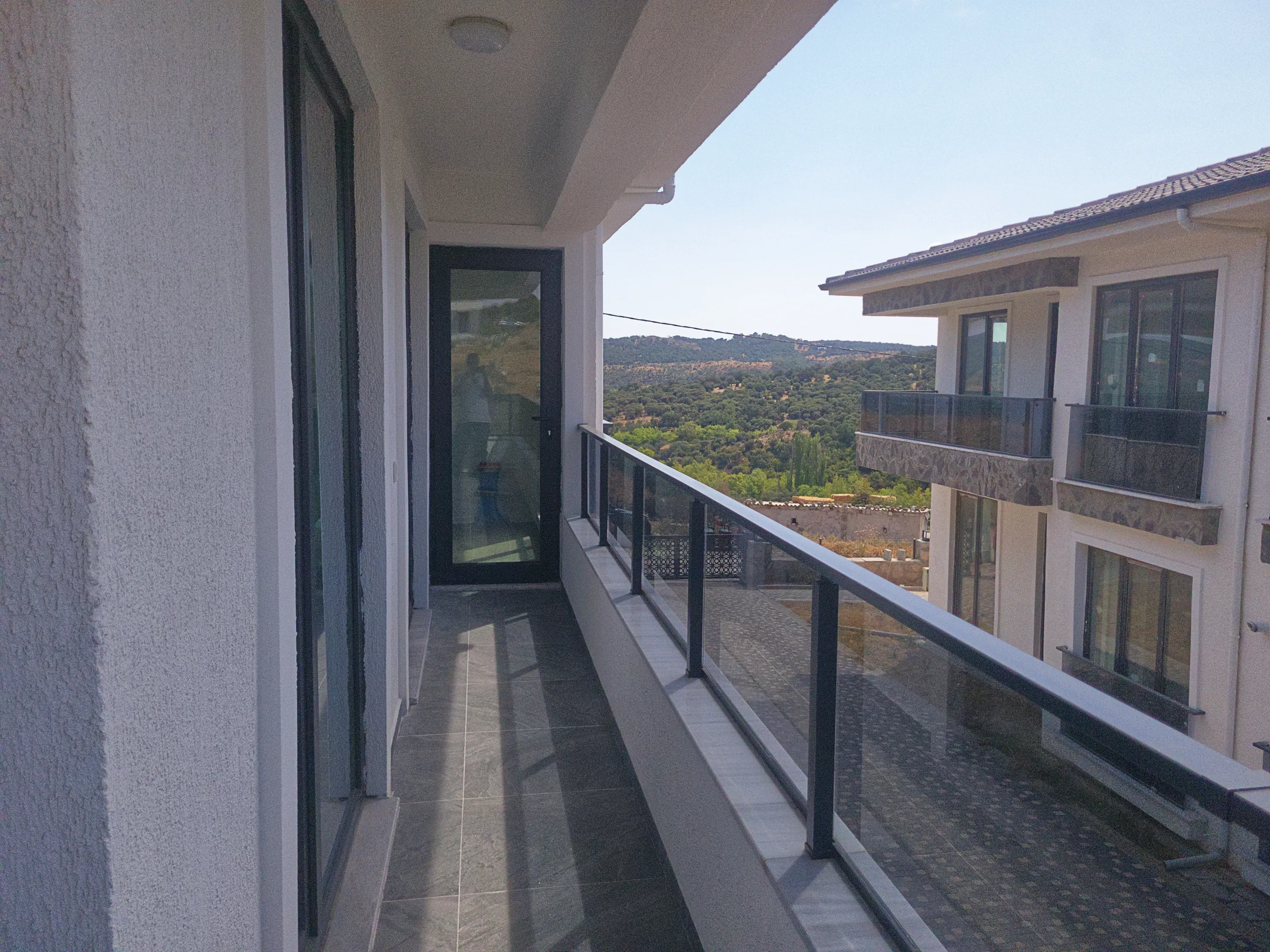 Long balcony with glass railings overlooking green hills and a neighboring residential building.