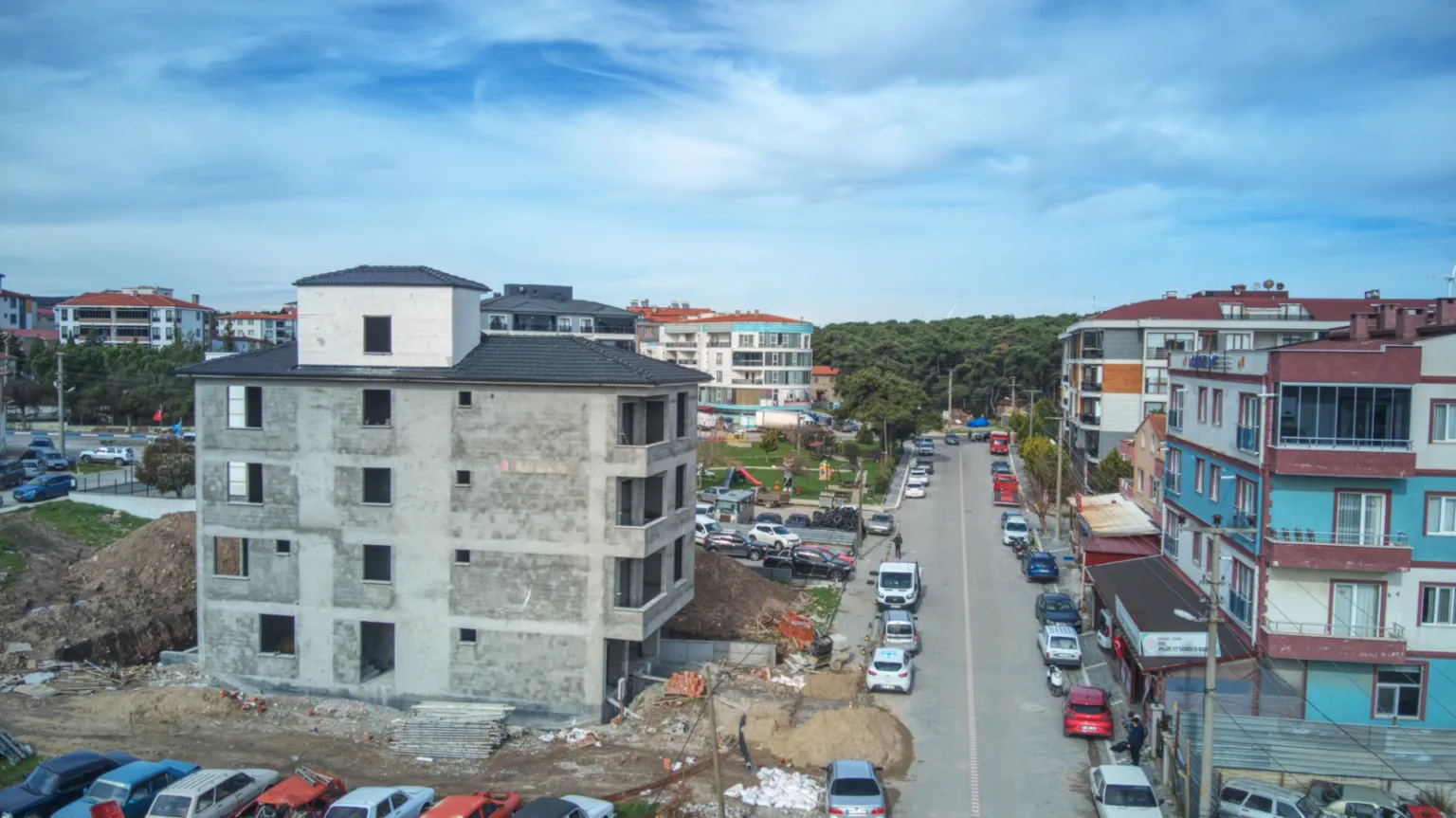 Elevated opposite-side view of the Small House project under construction, the adjacent street and surrounding buildings in Ayvacık.