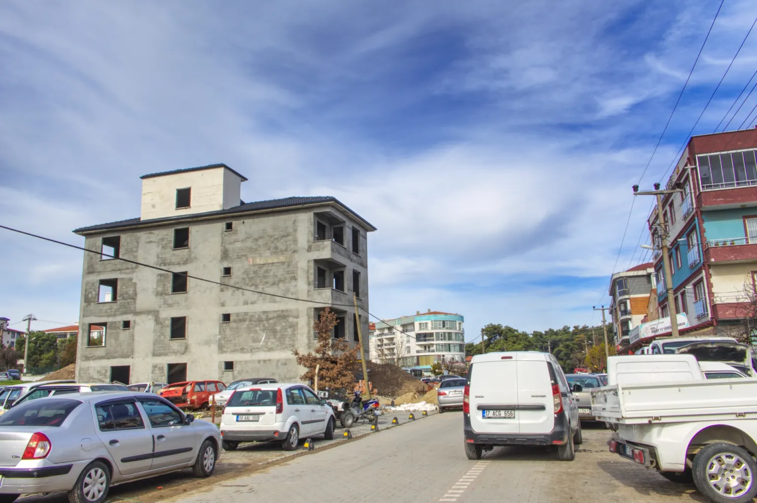 Small House projesinin tamamlanmamış beton cephesini ve önündeki araçlarla dolu caddeyi karşı köşeden gösteren görünüm.