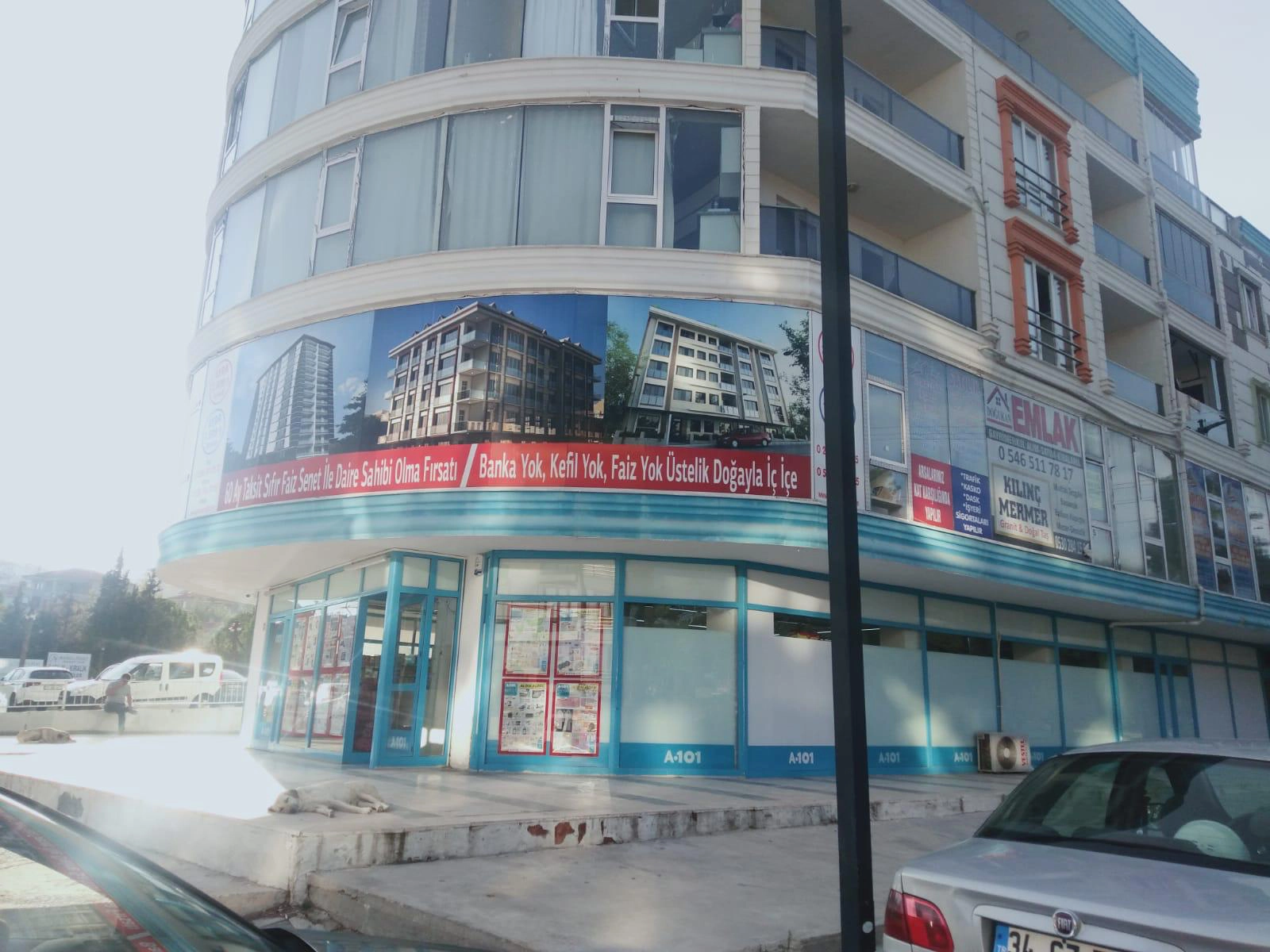 Çamlık Çarşı’nın kavisli köşe cephesi, zemin kattaki ticari alanları, üst kat balkonları ve dış cephedeki proje tanıtım afişi.