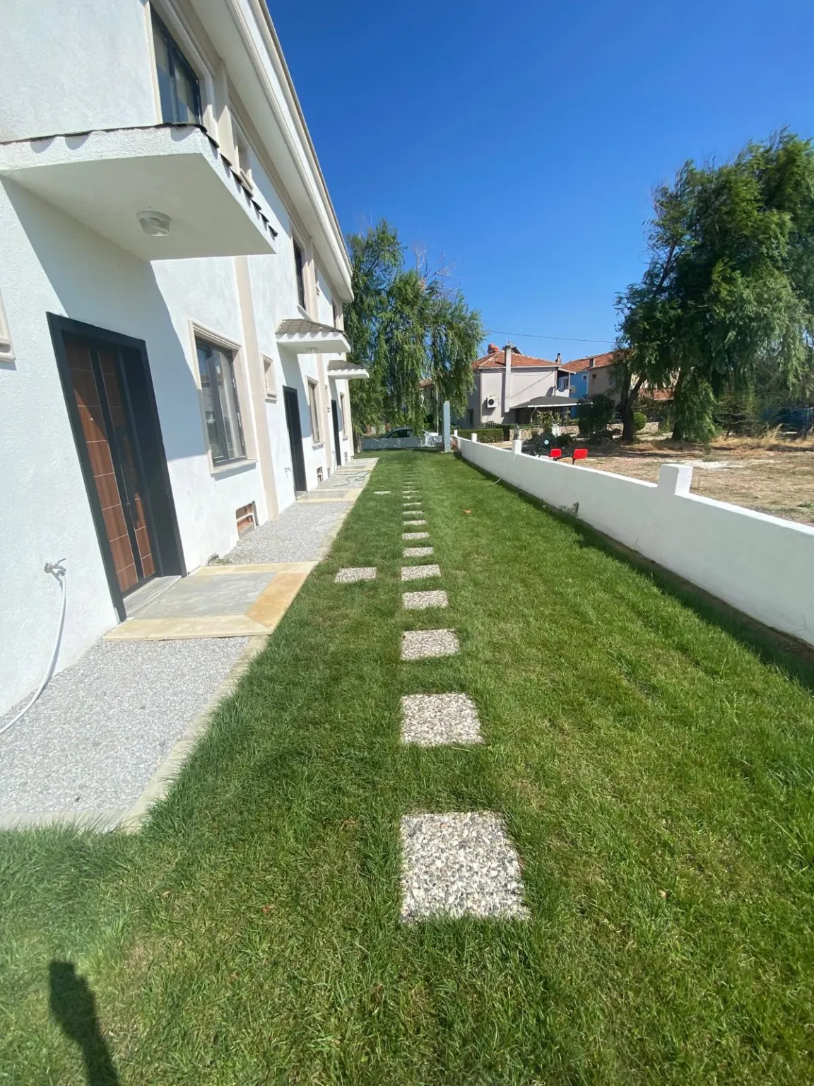 Beyaz konut ile bahçe duvarı arasında uzanan çim alan ve taş yürüyüş yolu.