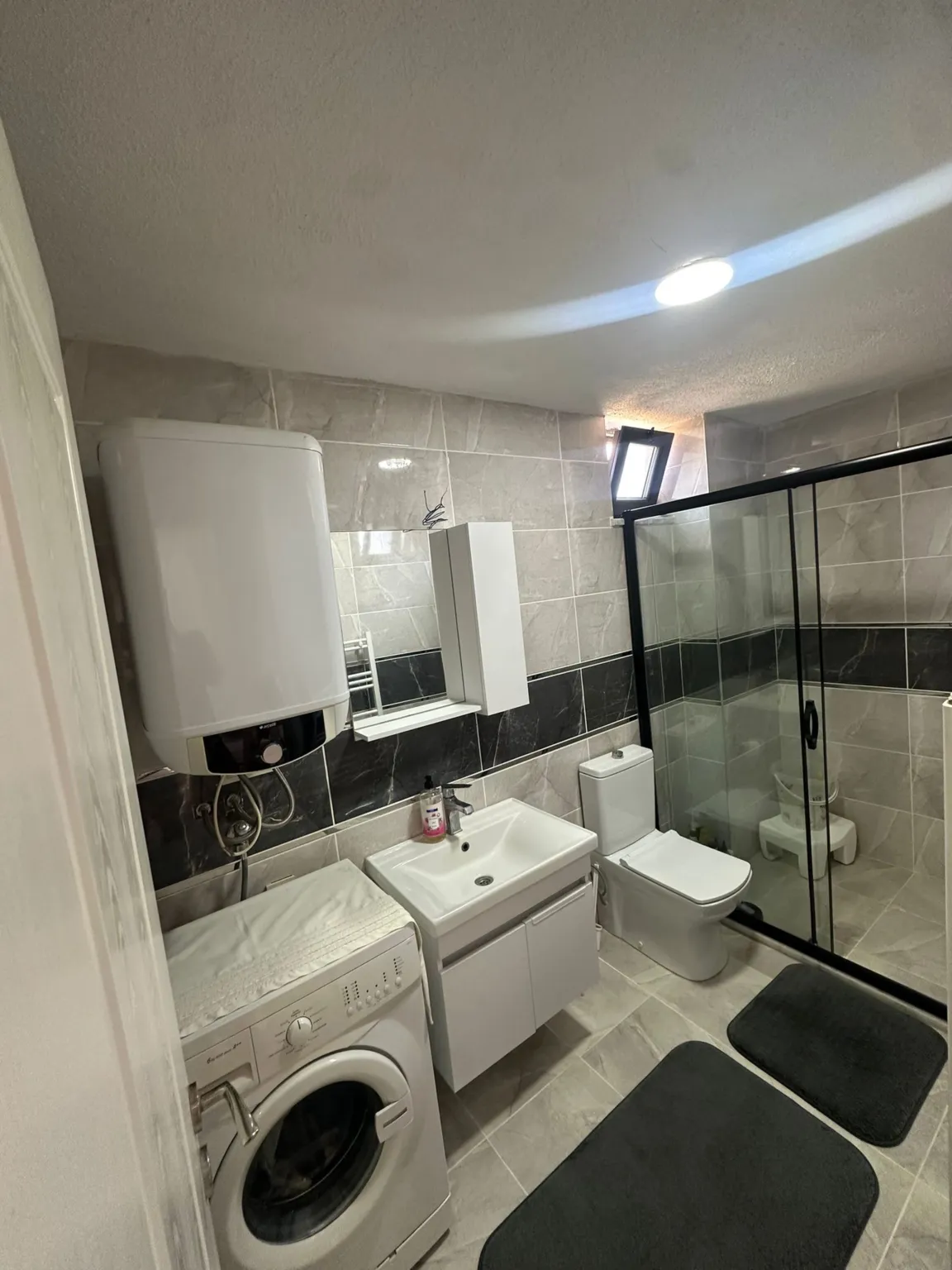 Cam duşakabin, lavabo, klozet, çamaşır makinesi ve su ısıtıcısı bulunan gri fayanslı banyo.