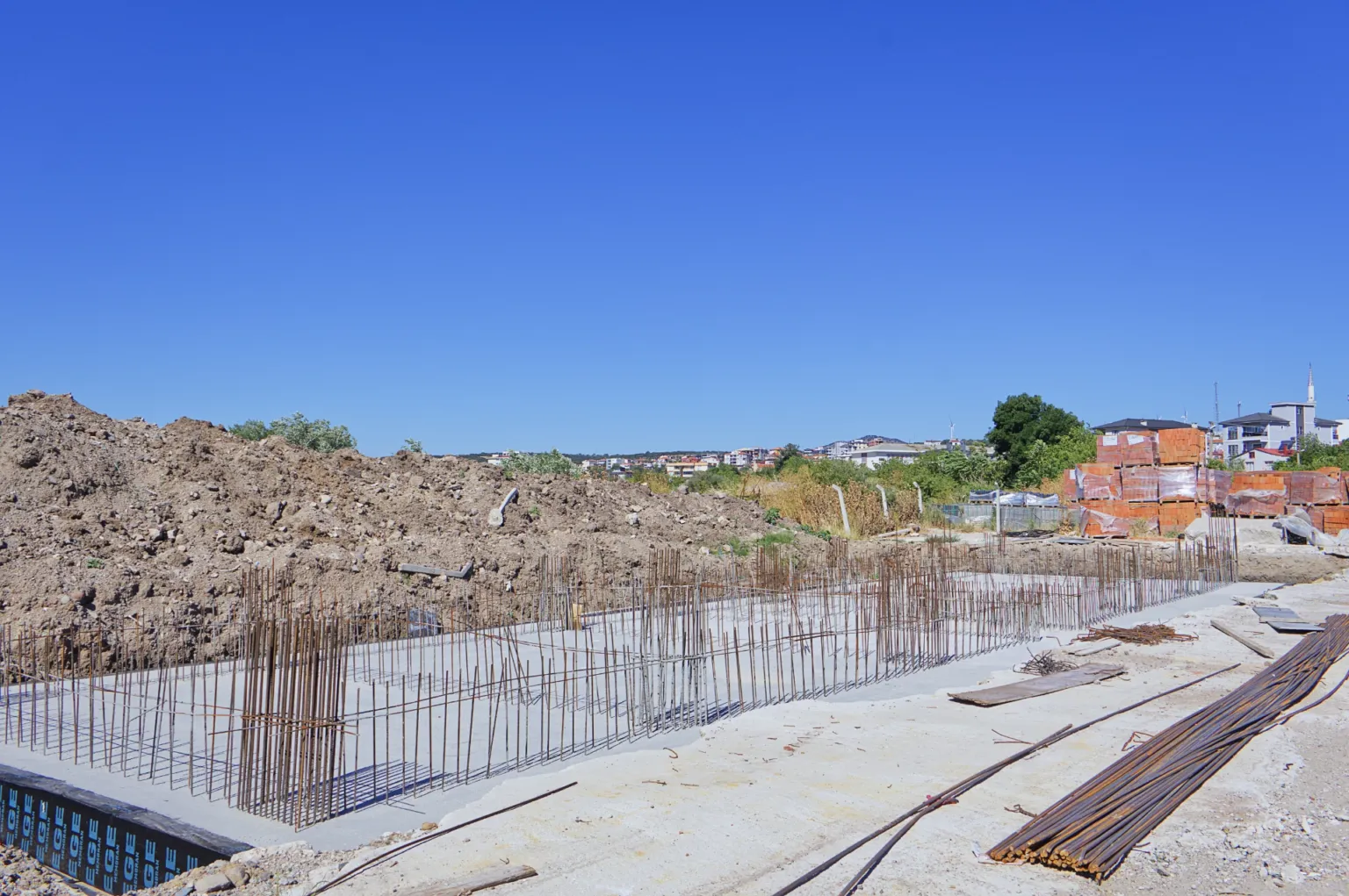 Side view showing steel reinforcement installed along the concrete foundation, stacked rebar, and piles of excavated soil.