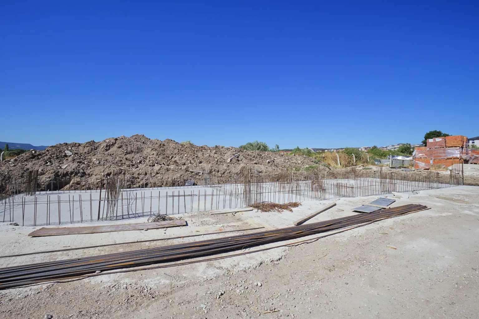 Beton temel tabanını, kenarlardaki dikey donatıları ve şantiye zemininde istiflenen çelik çubukları gösteren geniş açı.