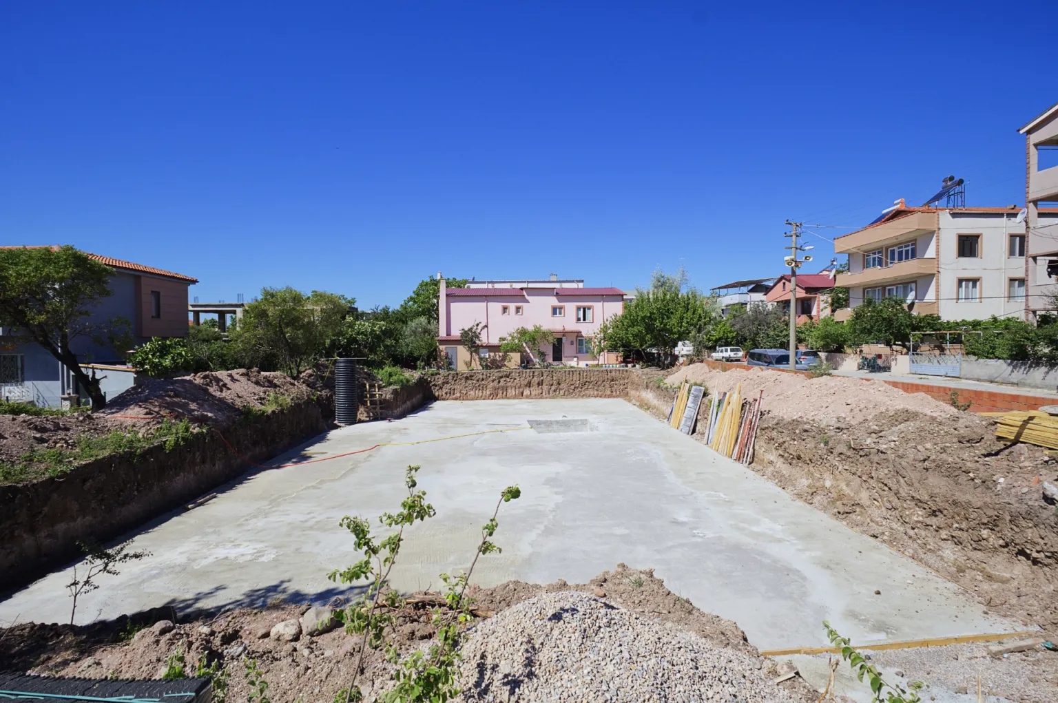 Çevresinde toprak yığınları ve kalıp malzemeleri bulunan beton temel tabanının geniş açıdan görünümü.