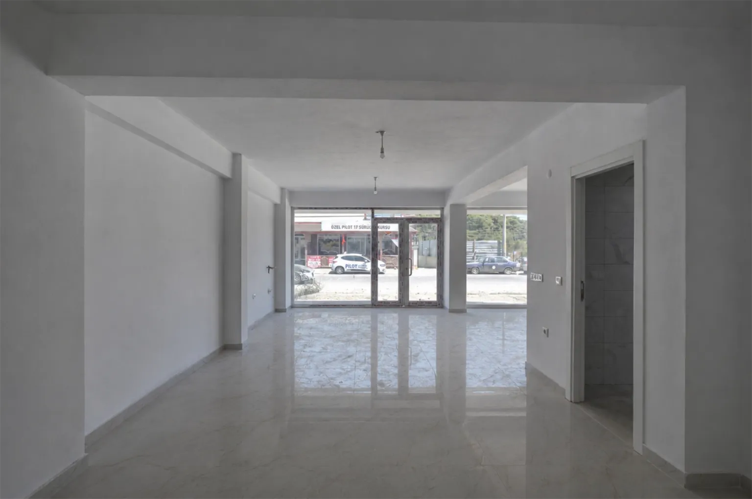 Street-facing commercial interior with white walls, a wide glazed entrance, glossy tile flooring, and a tiled side room on the right.