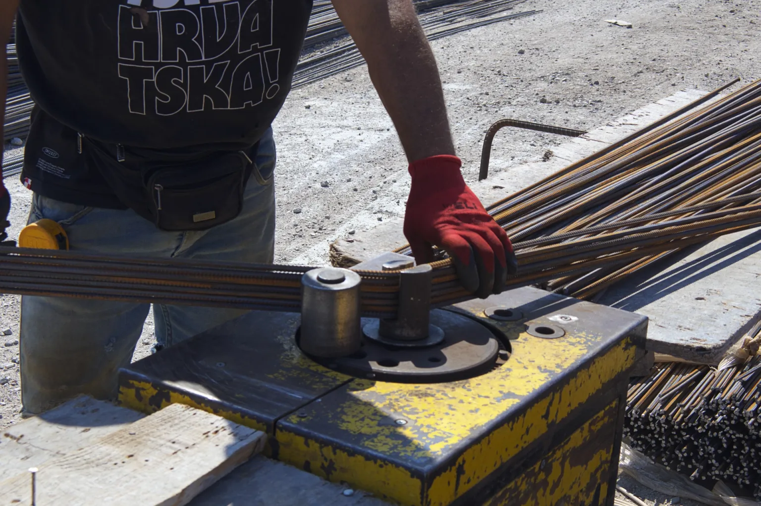 Close-up of a construction worker wearing red gloves and using a machine to bend a steel reinforcement bar.