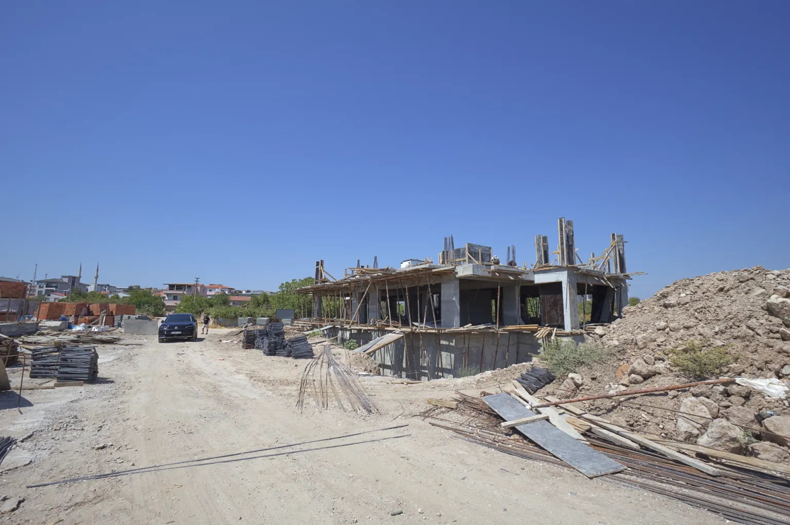 Toprak yol, istiflenmiş yapı malzemeleri ve hafriyat yığınları arasındaki betonarme yapının geniş şantiye görünümü.