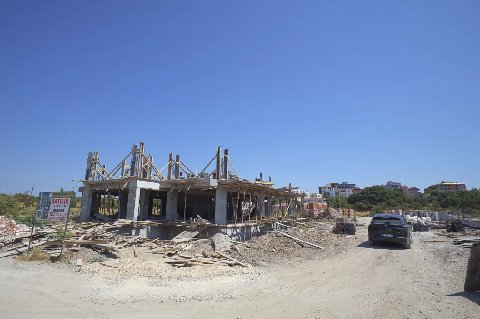 General view of a reinforced-concrete construction site with rising column reinforcement, building materials, and a car parked in front.