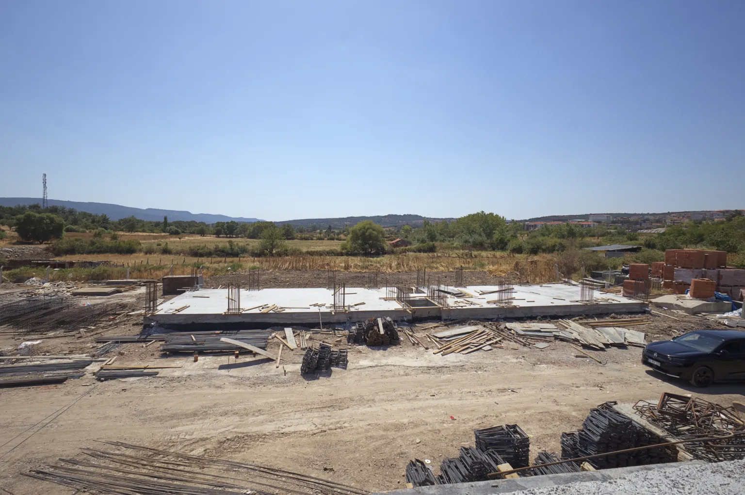 General view of the construction site showing the concrete foundation base, rising reinforcement, stacked building materials, and the surrounding rural landscape.