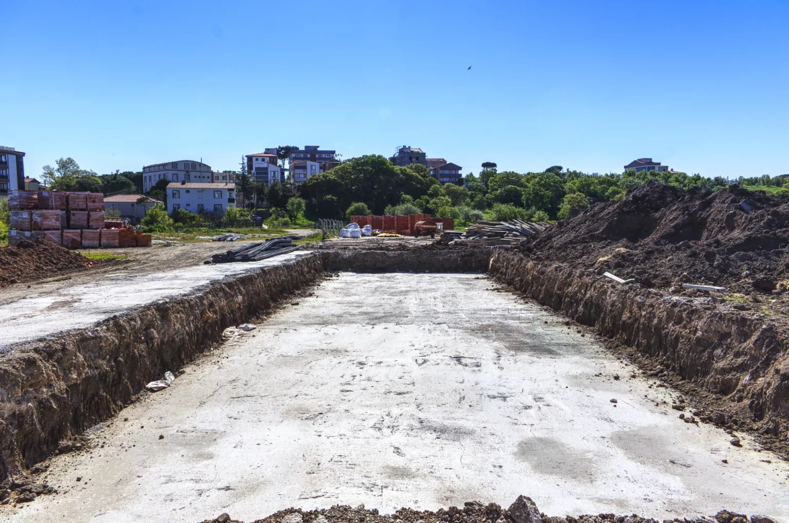 Kazı alanı içinde hazırlanan uzun dikdörtgen beton temel tabanını ve çevredeki toprak yığınlarını gösteren şantiye fotoğrafı.
