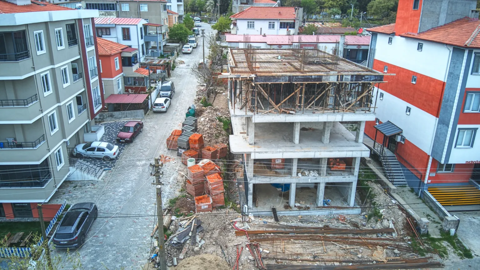 Cadde boyunca uzanan çevre yapılarının arasında yükselen Ezine Apartmanı şantiyesini ve sahadaki tuğla yığınlarını gösteren hava fotoğrafı.