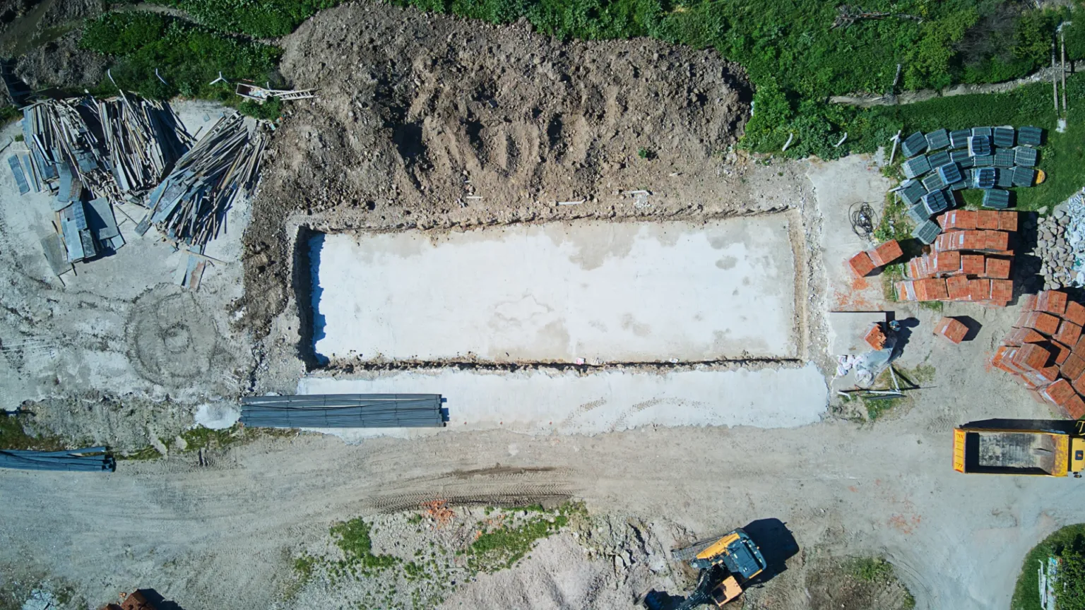 Top-down drone photo showing the concrete foundation base, surrounding excavation, construction materials, and machinery.