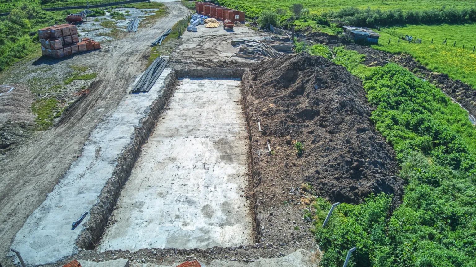 Yeşil arazinin yanında uzanan beton temel tabanını, kazılmış toprağı ve şantiye yolunu gösteren çapraz açılı hava fotoğrafı.