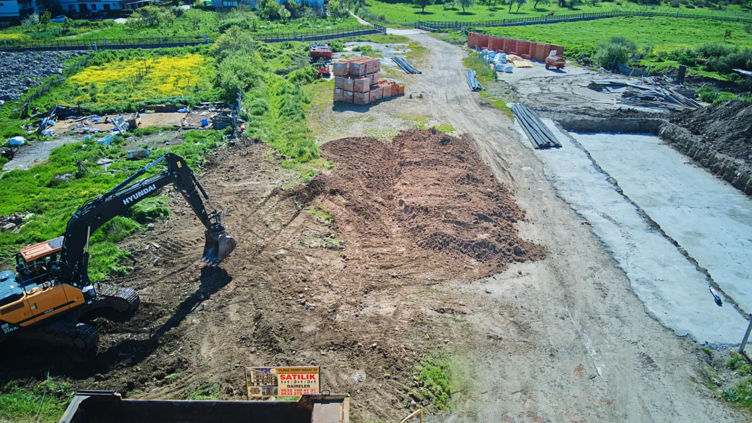 Aerial view of a construction site showing an excavator, earthworks, building materials, and a completed concrete foundation base on the right.