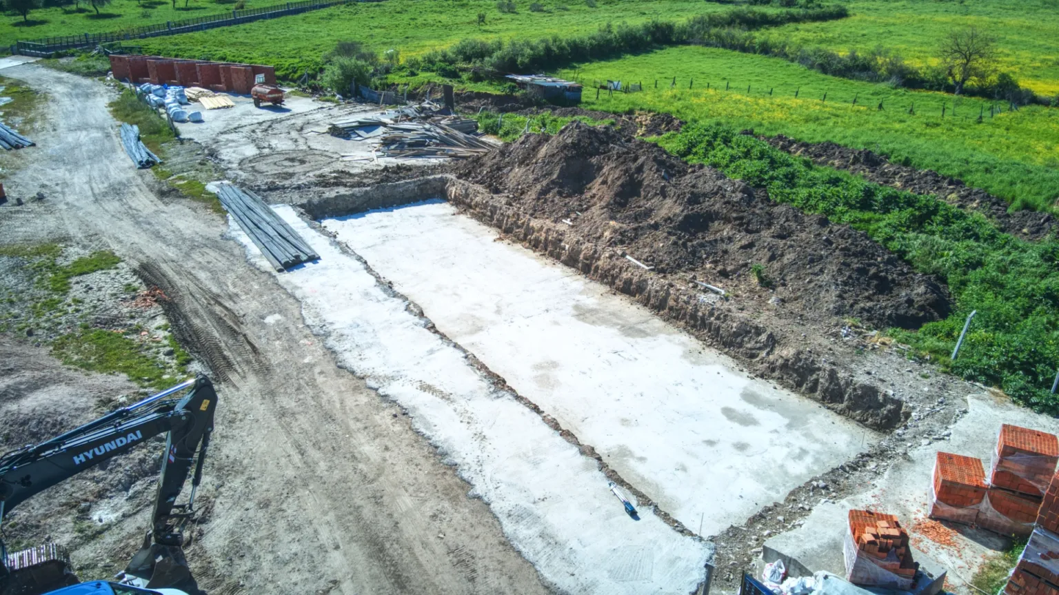 Temel hazırlık alanını, beton tabanı, toprak yığınlarını, tuğla paletlerini ve iş makinesini gösteren hava fotoğrafı.