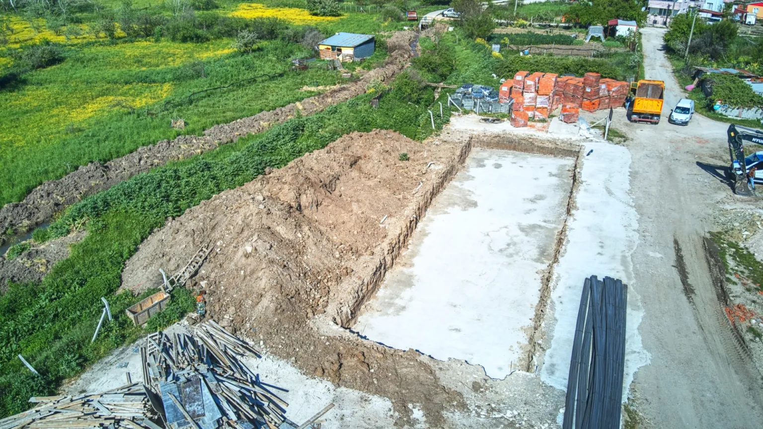 Beton temel tabanını, kazı sınırlarını ve çevrede istiflenen çelik profiller ile ahşap malzemeleri gösteren yüksek açılı şantiye fotoğrafı.