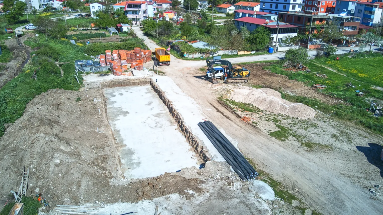 Temel tabanını, yanında istiflenen çelik malzemeleri, tuğla paletlerini ve şantiyedeki iş makinelerini gösteren drone fotoğrafı.