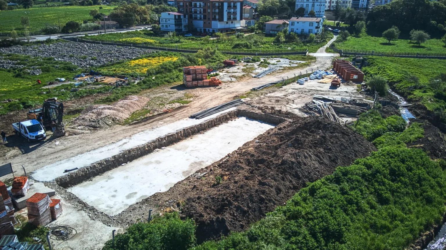 Yeşil tarlalar arasında yer alan temel kazısını, beton tabanı, toprak yığınlarını ve uzaktaki yerleşim alanını gösteren geniş hava fotoğrafı.