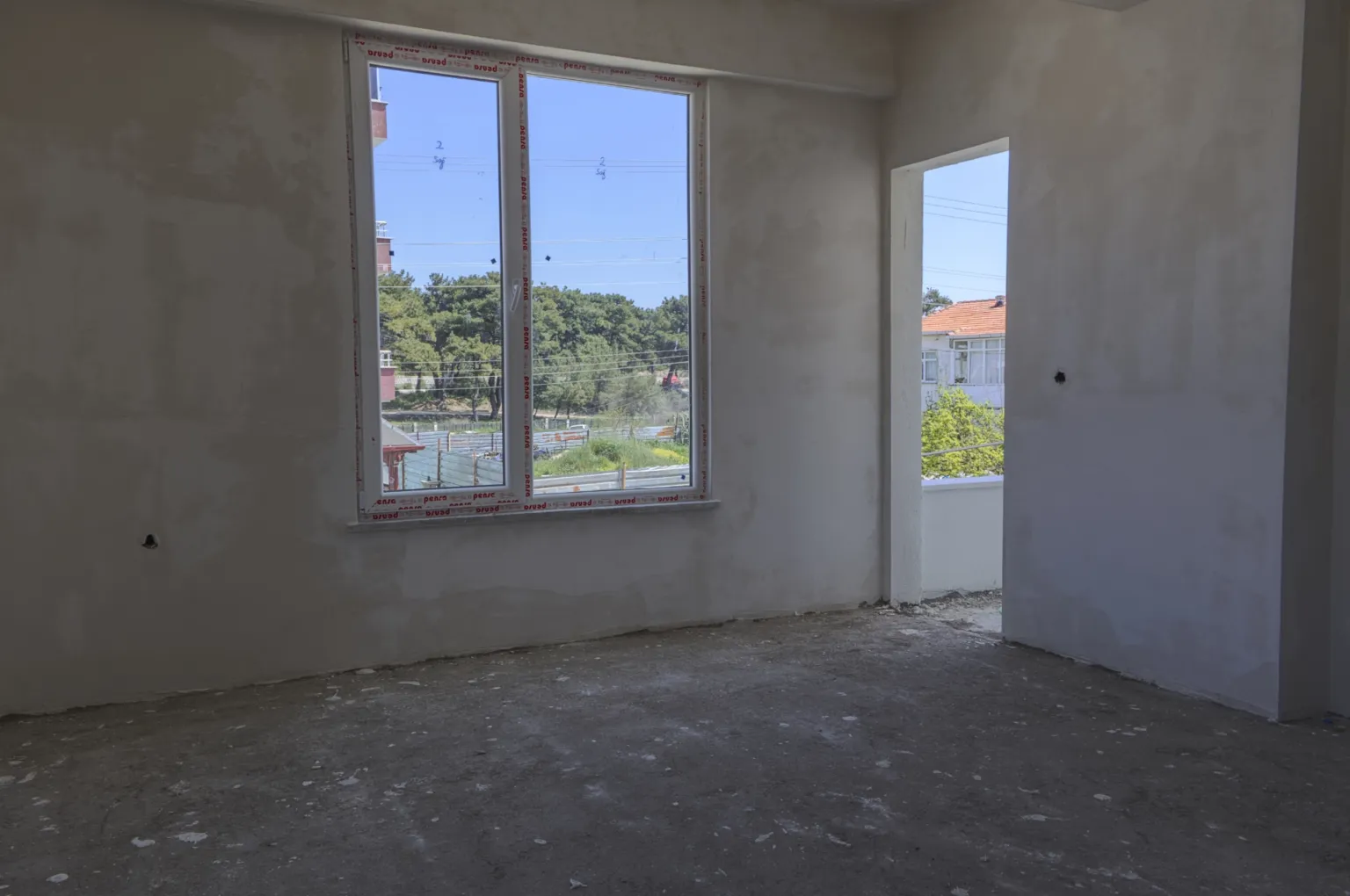 Small House projesinde pencere ve balkona açılan kapı boşluğuna sahip, sıvalı duvarlı ve ham beton zeminli oda.