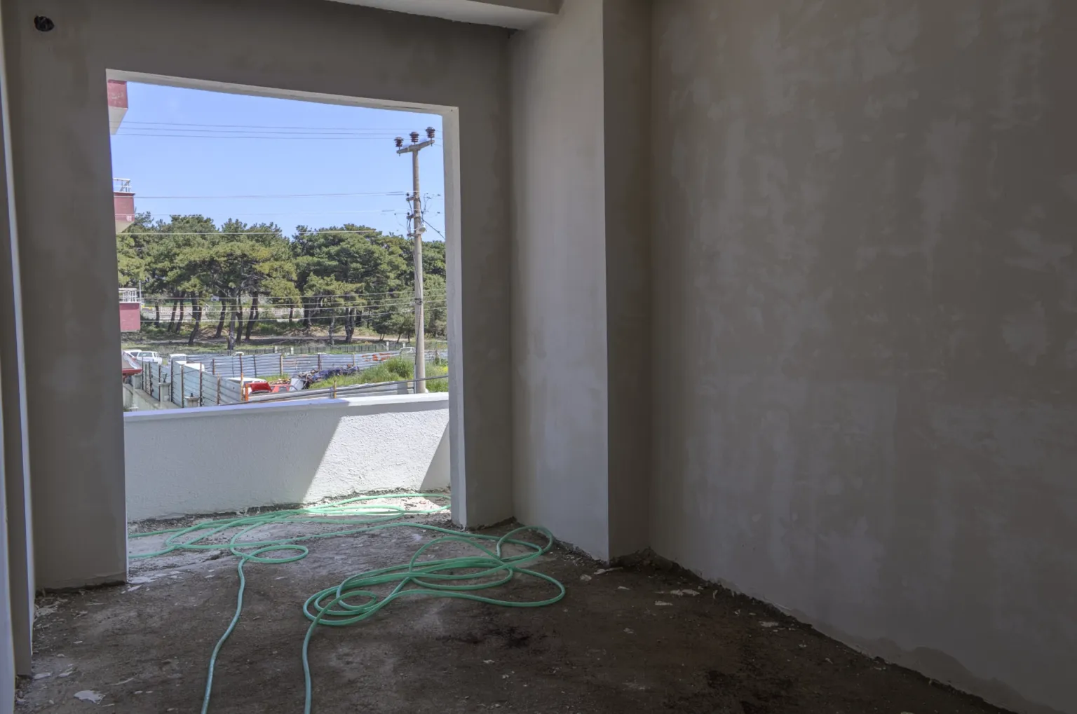 Unfinished room in the Small House project opening onto a balcony overlooking a wooded area, with plastered walls and a raw concrete floor.