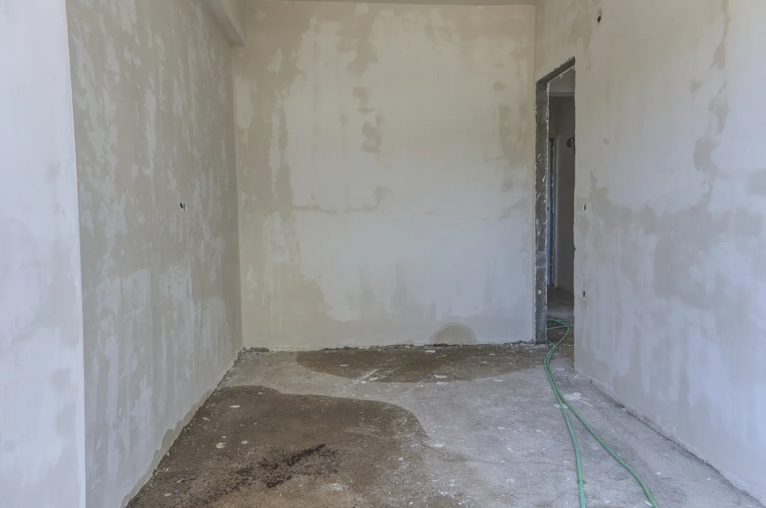 Unfinished interior in the Small House project with plastered walls, an incomplete door frame, a raw concrete floor and green hose.