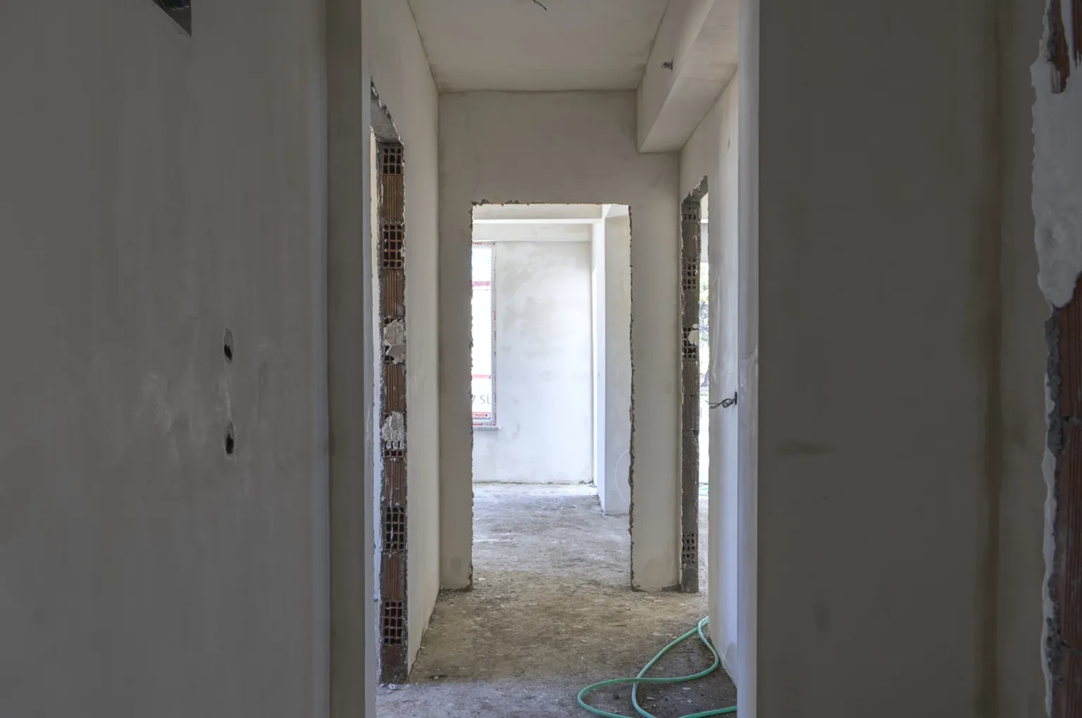 Small House projesinde sıvalı duvarları, tamamlanmamış kapı açıklıklarını, ham beton zemini ve yeşil hortumu bulunan iç koridor.