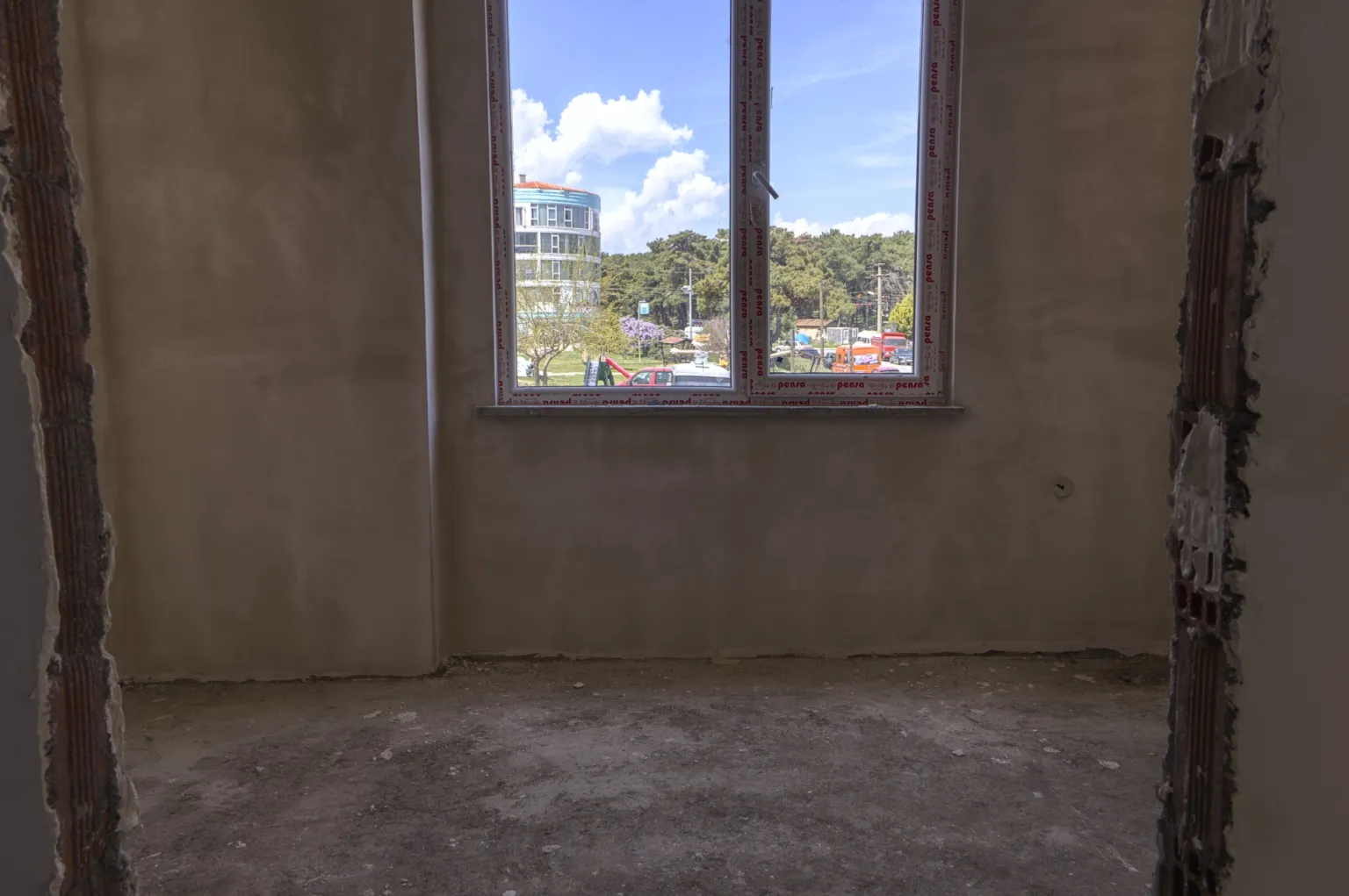 Small House projesinde ham beton zeminli, sıvalı duvarlı ve kent manzarasına açılan pencereli tamamlanmamış oda.
