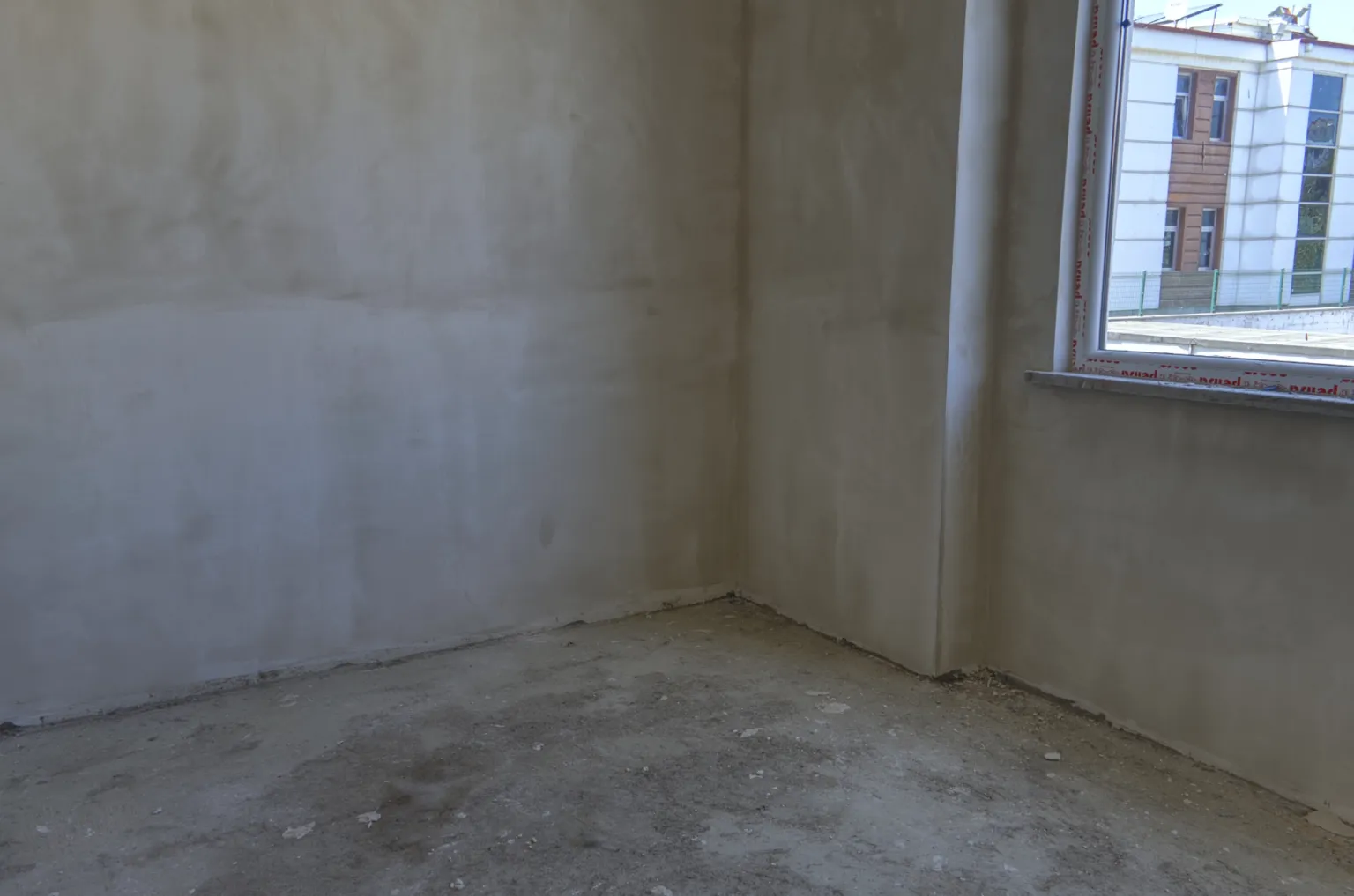 Small House projesinde sıvalı duvarları, ham beton zemini ve karşı binaya bakan penceresi bulunan boş oda.