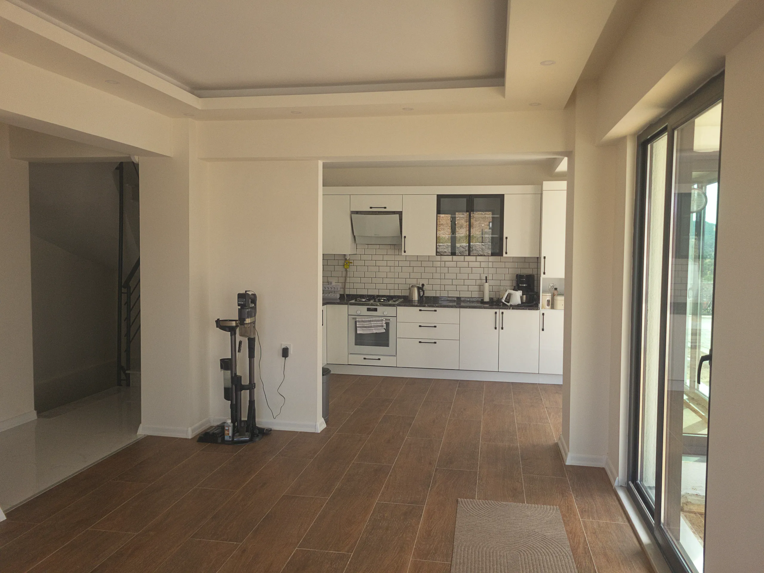 Modern residential interior featuring wood-look flooring, a white open-plan kitchen, and large glass doors.