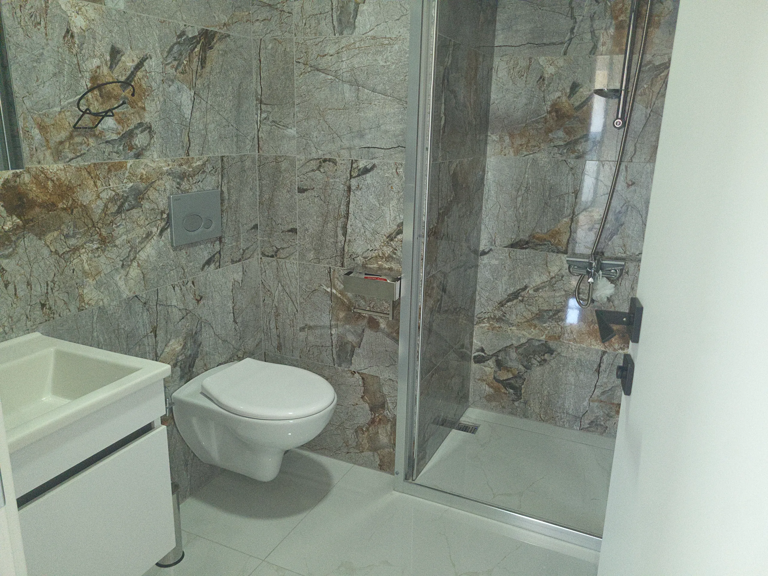 Modern bathroom with marble-look wall tiles, a glass-enclosed shower, wall-mounted toilet, and white vanity.